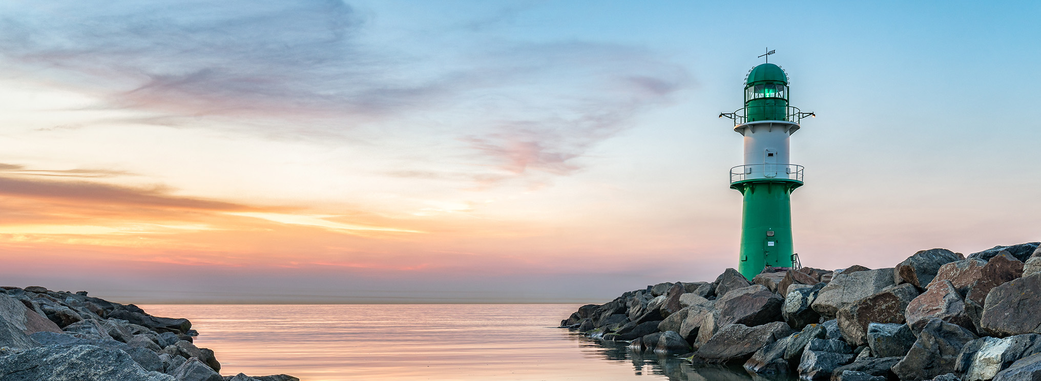 Nach dem Sonnenuntergang liegt sanftes Licht über der steinigen Küste von Warnemünde. Das ruhige Meer und der Leuchtturm schaffen einen stillen Ort des Innehaltens und Erinnerns.