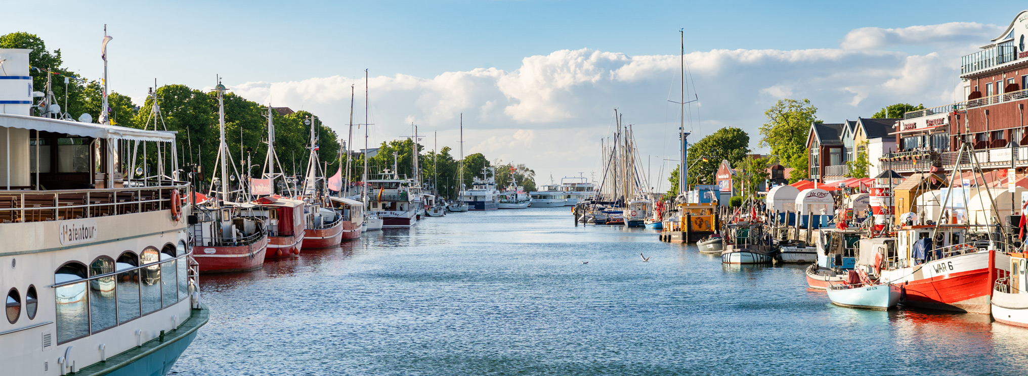 Der Hafen von Warnemünde zeigt sich an einem sonnigen Tag mit ruhiger Atmosphäre. Die Szene verbindet maritimes Leben mit Gelassenheit und Weite.